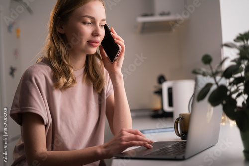 Concept of Freelance. Young woman in pajamas working on laptop and talking on smartphone at home kitchen