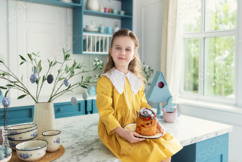 Wallpaper Mural Young girl sit on kitchen with cake, spring decor, flowers, easter eggs and bunny. Child Girl in yellow dress holding easter cake on plate in hands. Little Girl preparing festive pastries at home. Torontodigital.ca