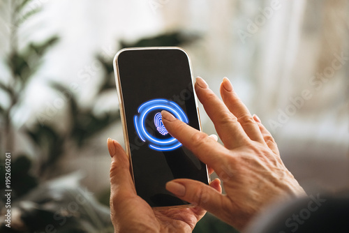 Close-up of a hand using fingerprint recognition on a smartphone screen with a blue fingerprint icon illuminated, surrounded by blurred indoor plants in the background