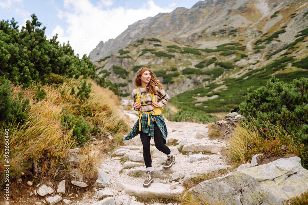 Obraz premium A female hiker with a backpack walks along a rocky hiking trail in the mountains. A smiling woman enjoys the mountain scenery at sunset. Concept of hiking and adventure.