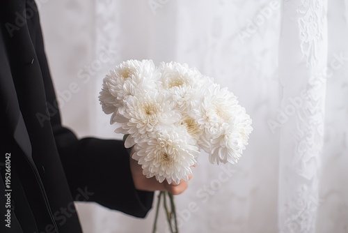 Hand holding a bouquet of white chrysanthemums against a soft white curtain backdrop, symbolizing purity, remembrance, or mourning, with a black sleeve adding contrast. 