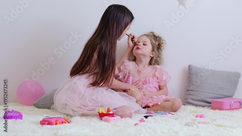 Two sisters are having fun as one applies makeup to the other. They sit on a soft rug in a well-lit room filled with toys and colors