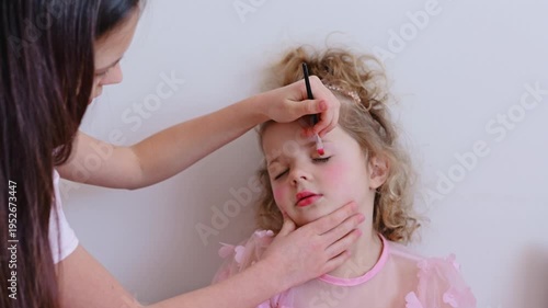 A young child sits while an older helper applies makeup on her face in a bright indoor space during daytime, showing a moment of fun and creativity