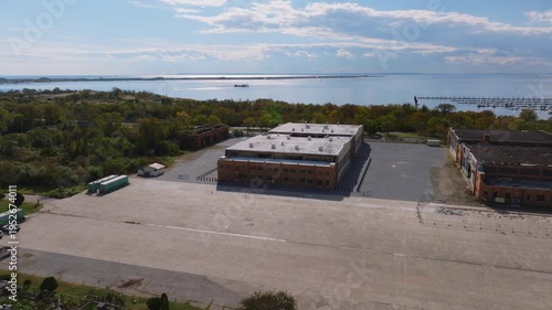 Aerial drone view of an industrial facility with warehouses, trucks, and equipment near a coastal landscape with water and greenery under a cloudy sky.