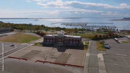 Aerial drone view of an industrial facility with warehouses, trucks, and equipment near a coastal landscape with water and greenery under a cloudy sky.