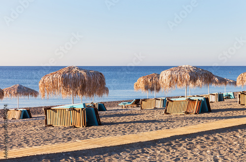 A Mediterranean sea sandy beach with straw umbrellas and folded sun loungers early in the sunny morning. Seascape, summer natural backdrop. Summer seaside vacation concept