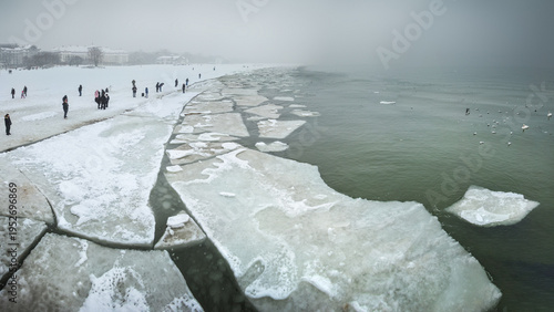 Winter landscape of a frozen Baltic Sea in Sopot, Poland, with ice floes on the water and people walking on the snowy beach under a foggy sky