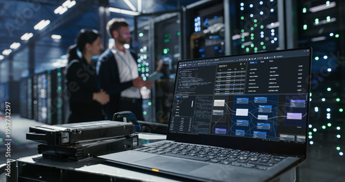 Laptop sits on a cart beside hard drives in data center.  Two people discuss network infrastructure and server maintenance among  server racks and mainframes.