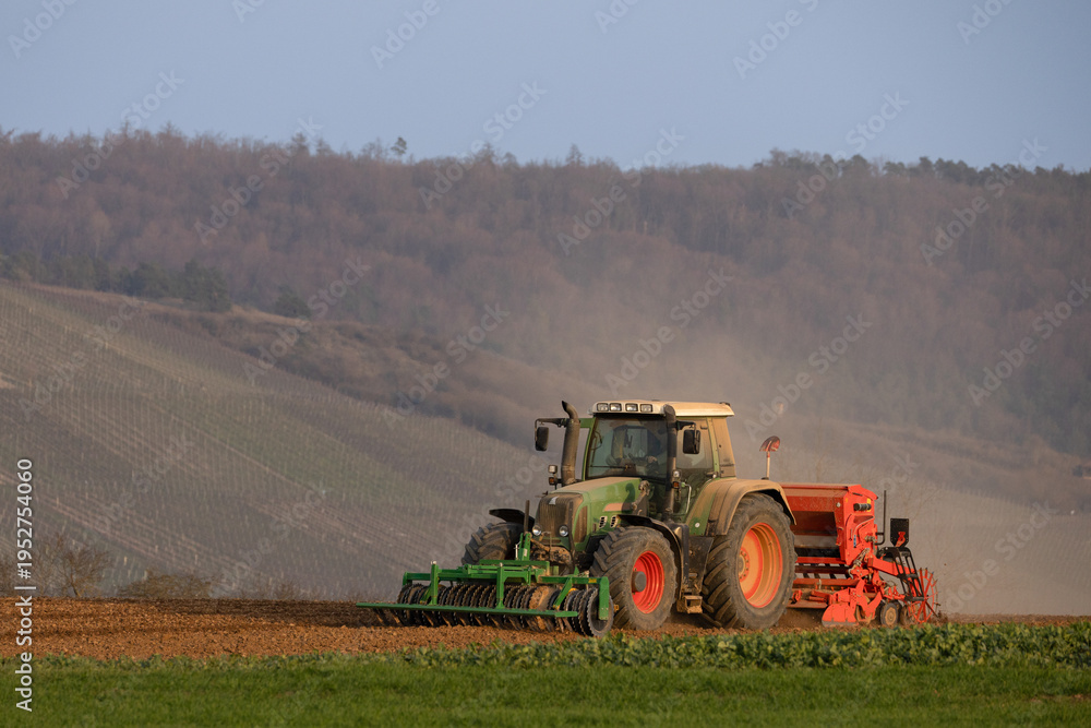 Fototapeta premium Tractor working on agricultural field with seeding machine in rural landscape at sunset