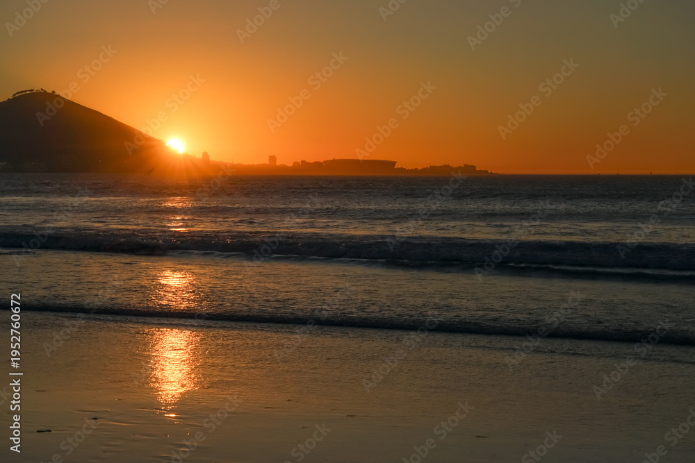 Obraz premium South Africa,Cape Town,Sunset over a calm ocean with waves gently rolling onto the beach, reflecting golden light.
