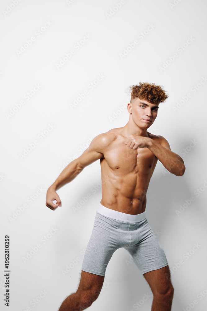 Fototapeta premium Muscular young man in light grey shorts with defined abs posing with arms raised on white background. Athletic body, youthful fitness, curly hair and confident expression, isolated studio portrait.