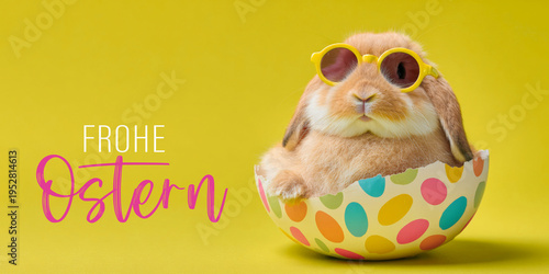 Frohe Ostern Konzept, Ferien Feiertag Grußkarte, Karte mit deutschem Text - Lustiger Osterhase mit Sonnenbrille sitzt in einem buntem Osterei isoliert auf gelbem Hintergrund