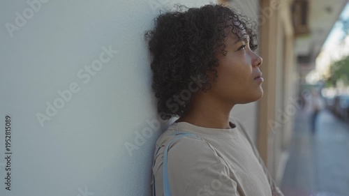 Woman leaning against wall on city sidewalk with face turned upward and eyes looking skyward; quiet reflection.