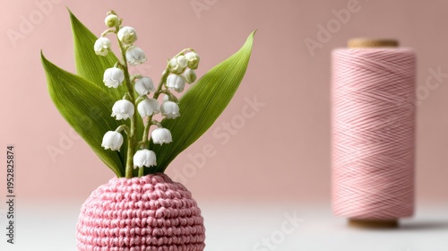 Pink crocheted vase holds white lily of the valley flowers. Green leaves add fresh contrast. Soft pink thread spool sits in background. Gentle, springtime, handmade craft theme
