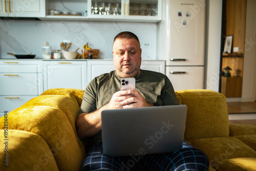Middle-aged man using smartphone and laptop on couch at home