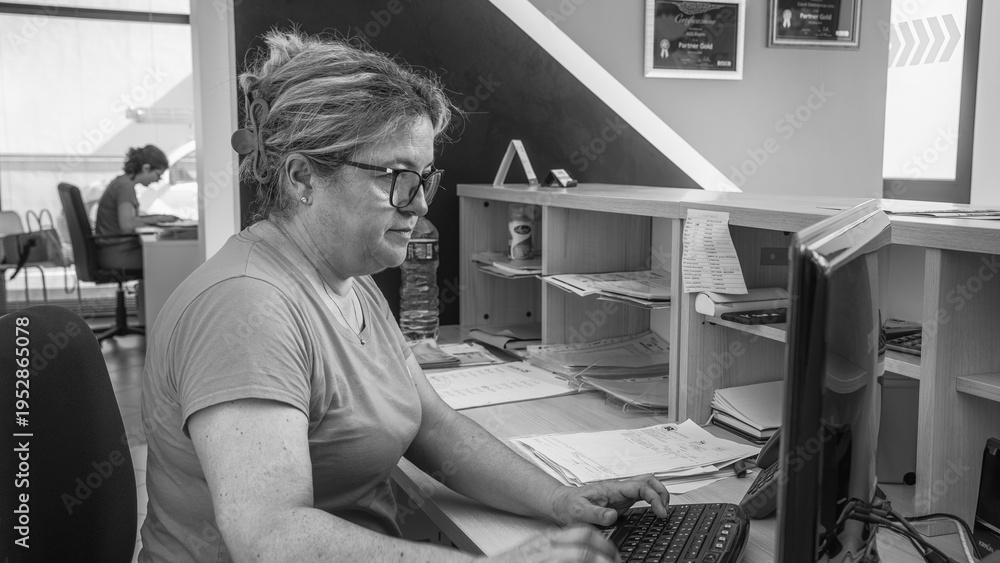 Fototapeta premium Smiling female secretary working efficiently at her office desk using a desktop computer and phone