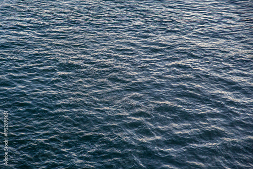Top-down view of deep blue sea water with gentle ripples and soft light reflections. The surface shows a rhythmic pattern of small waves, capturing the vast and calm essence of the open ocean.
