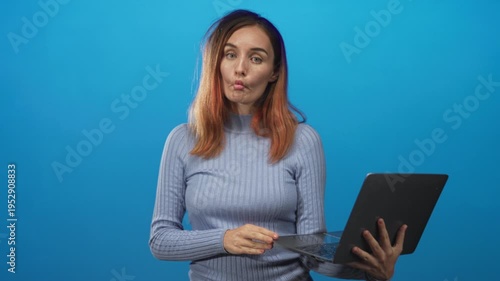 Young woman with red hair holding laptop, fingers on trackpad, puckering lips, wearing light blue sweater in studio; playful focus.