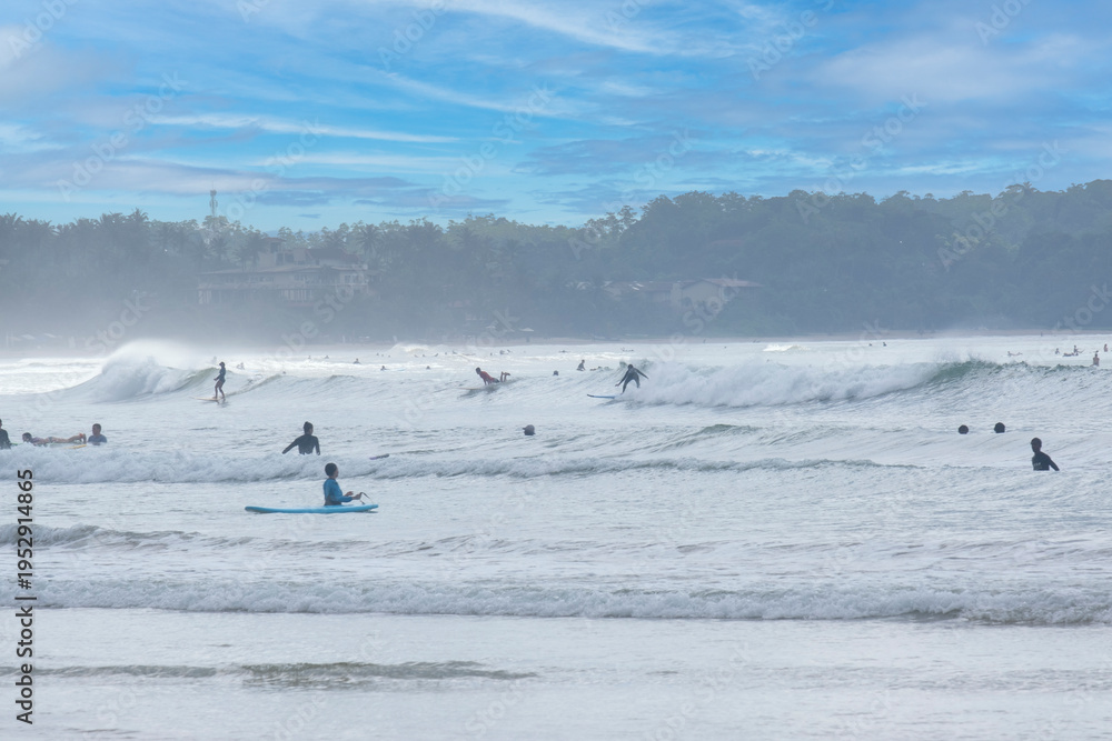 Naklejka premium 8.12.2025. Weligama Sri Lanka. Crowded surf spot in Weligama bay, Sri Lanka. Group of young people and beginners learning to surf with boards on the beach and in the Indian ocean waves.