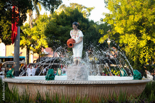 Valladolid, Mexico; 1st November 2025: Traditional Mexican woman statue holding a clay pot in a splashing fountain surrounded by lush greenery.