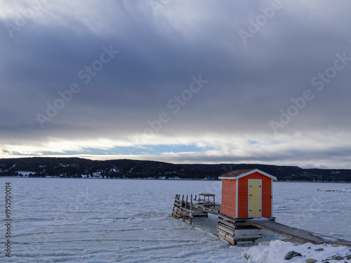 An orange fishing shed stands at the middle of a wooden dock overlooking a vast, frozen ocean beneath heavy winter clouds.