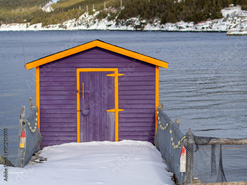 A brightly painted purple shed with yellow trim stands on a snowy dock overlooking calm winter water.