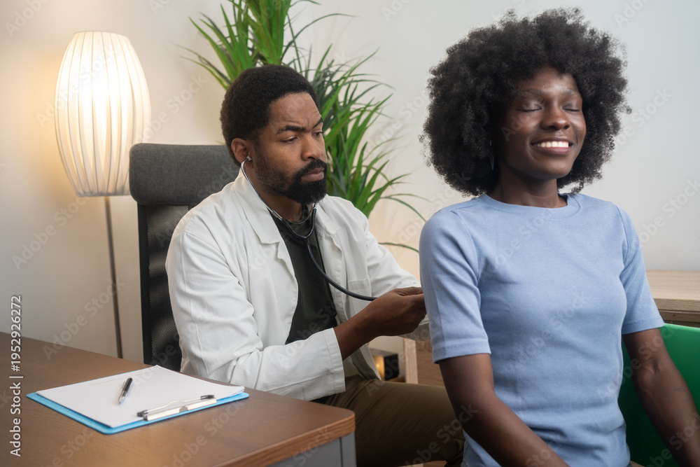 Fototapeta premium Black male doctor listening to female patient’s lungs with stethoscope in clinic