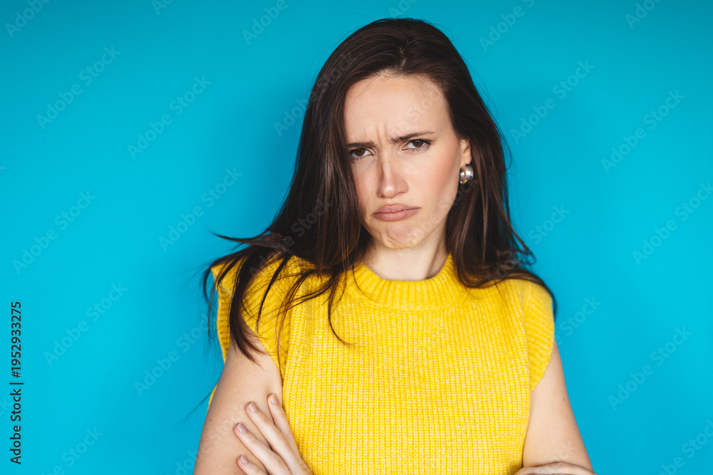 Fototapeta premium Offended and grumpy young woman with crossed arms looking at camera with skepticism and dislike, feeling annoyed or bored, standing against blue background in studio portrait