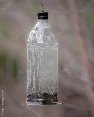 A repurposed plastic bottle hangs by a wire, filled with seeds and fitted with a small perch for visiting birds.