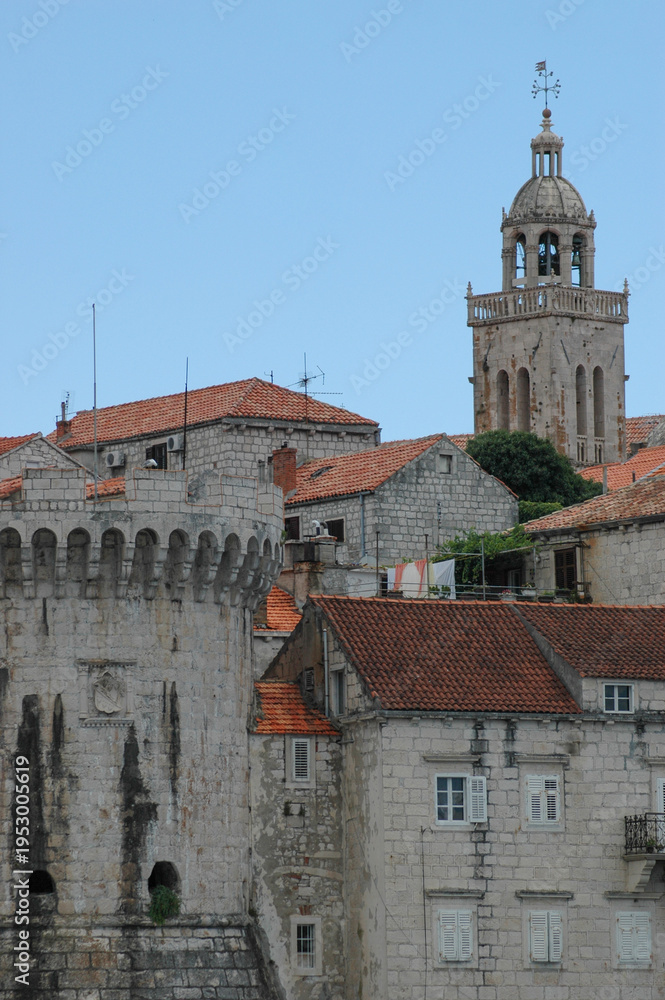 Obraz premium A scenic view of the historic Korcula old town featuring a massive stone fortification tower and the bell tower of St Mark Cathedral rising above red terracotta roofs