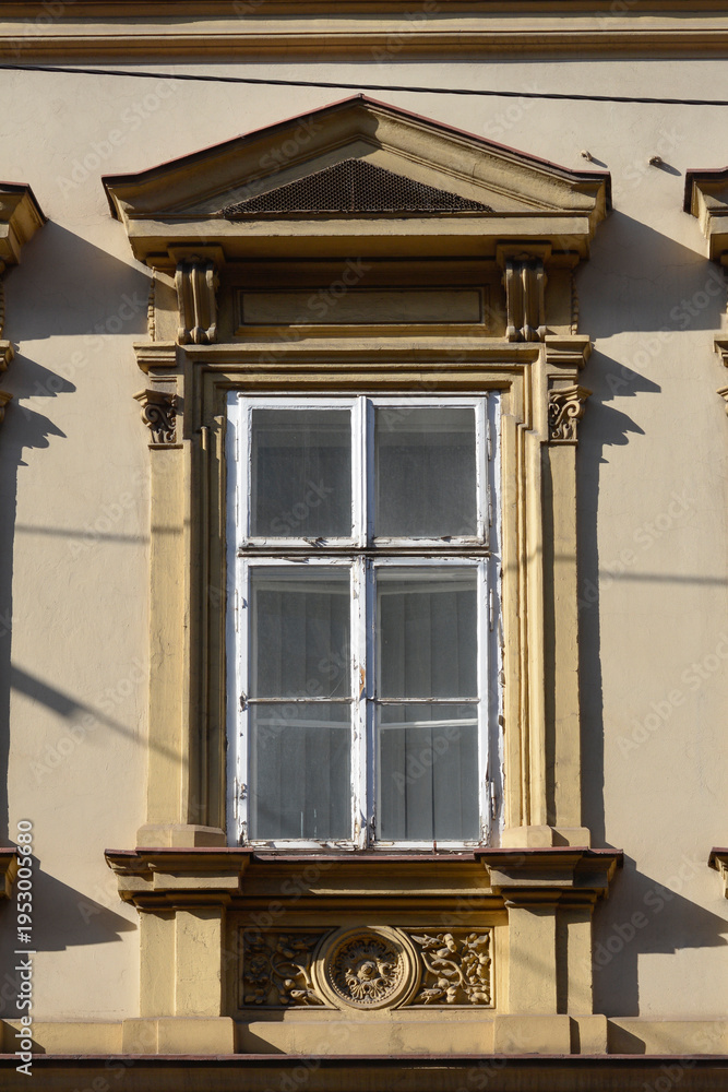 Obraz premium An old white wooden window featuring a decorative stone frame with classical ornaments and a triangular pediment on a beige building wall