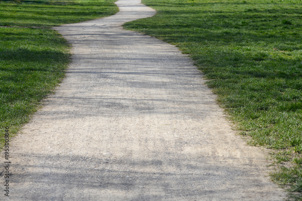 Obraz premium A light colored winding gravel walkway curves through a lush green field of grass in a sunny outdoor park