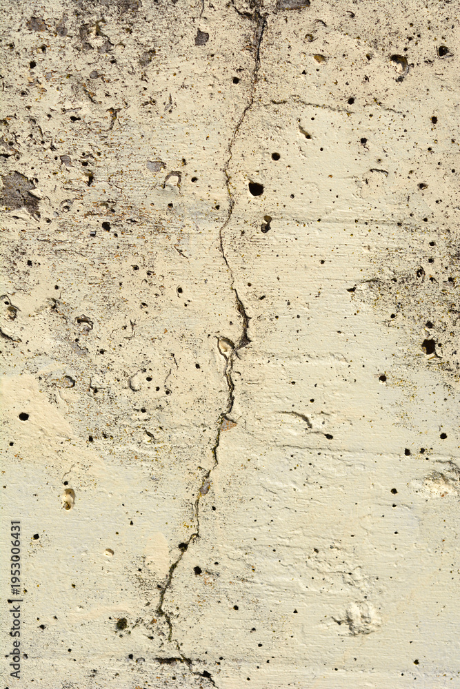 Obraz premium A full frame vertical shot of a weathered beige concrete wall featuring a prominent jagged vertical crack and a porous pitted surface