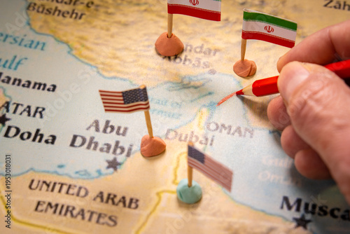 Conceptual image of a hand drawing a red line on a map of the Middle East with United States and Iran flags marking the Strait of Hormuz. Represents geopolitical conflict and global oil supply crisis.