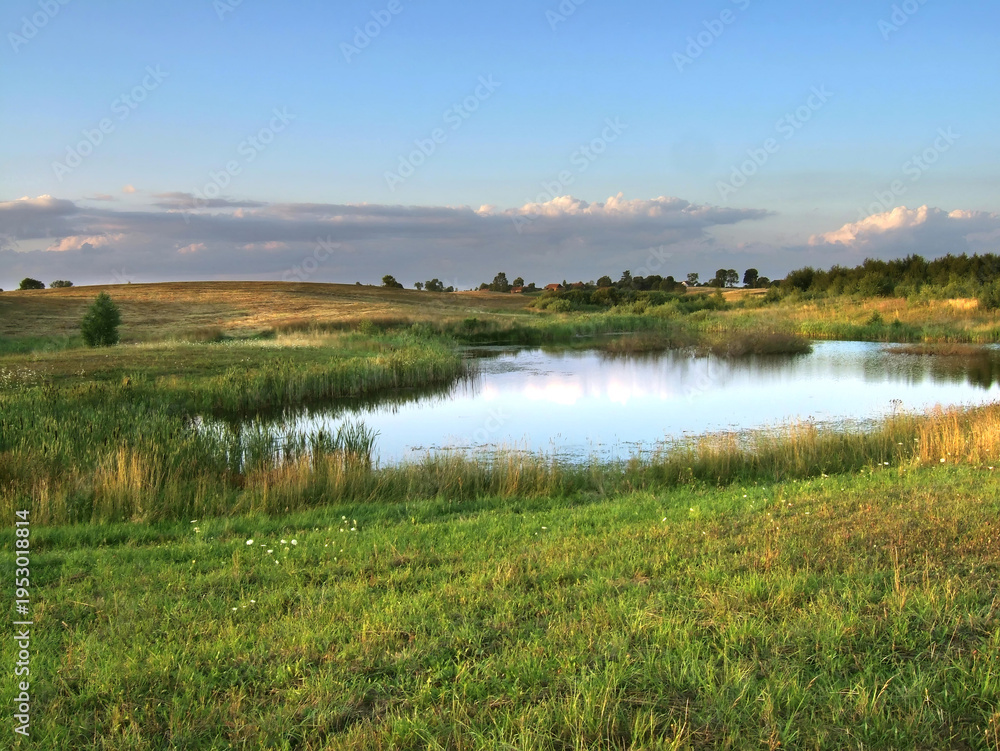 Fototapeta premium A lush green meadow and pond under a evening summer sky in the rural Masurian region. Distant forests create a scenic pastoral view of the Polish countryside.