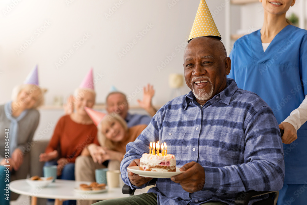 Naklejka premium A man in a wheelchair smiles while holding a birthday cake with candles. Friends and caregivers surround him, wearing party hats. The event takes place indoors in a bright room.