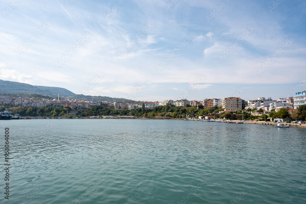 Fototapeta premium Scenic view of a Mediterranean coastal town with multi-story buildings - boats anchored in calm turquoise waters - and mountainous backdrop under partly cloudy skies