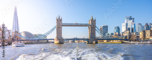 A ship moves on the river, showing Tower Bridge in London. Tall buildings fill the background under a clear sky. Water pushes away from the ship as it travels.