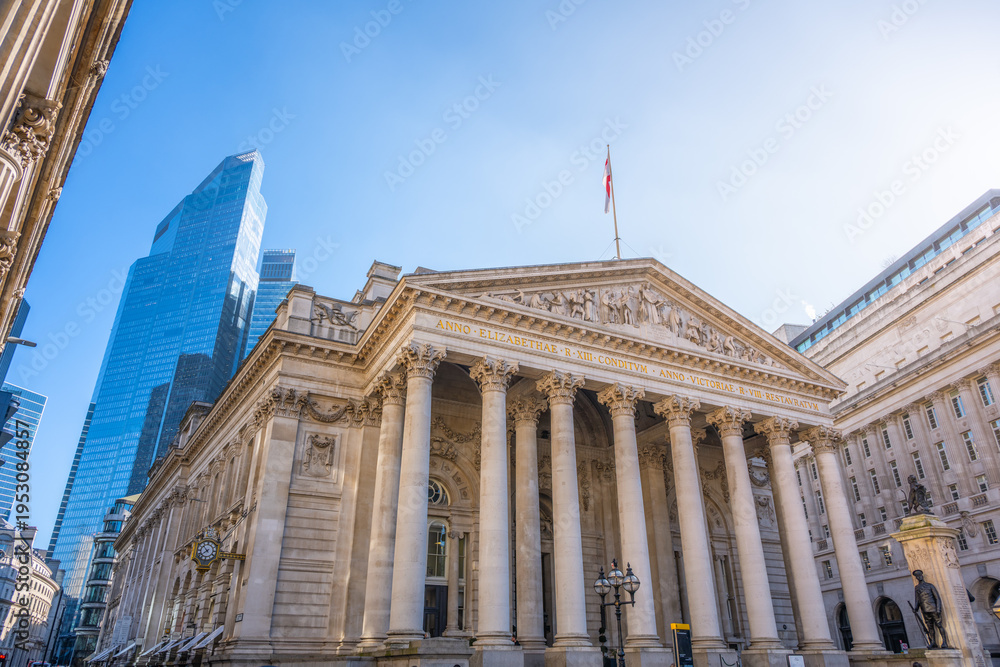 Fototapeta premium The Royal Exchange in London stands among modern structures on a clear day. People walk nearby, and the British flag flies high above the historic site.