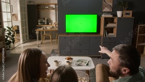 Rear zoom-in shot of cheerful Caucasian parents and young daughter enjoying fun comedy on smart TV with empty green screen, using subscription streaming service in cozy relaxing home interior