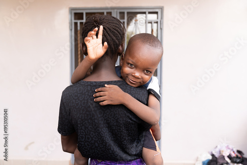 A small African boy, carried by his older sister, waves his hand joyfully – a symbol of familial care and sibling love.