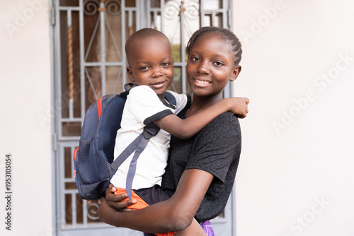 Skeptical little African boy on his first day of school with a backpack on his shoulders, in the arms of his older sister who tries to encourage him