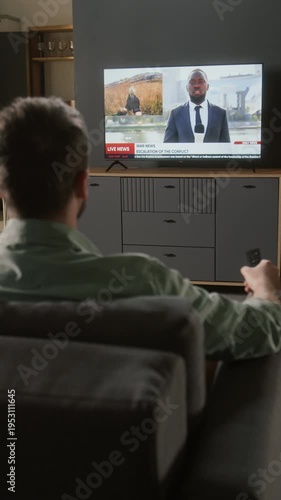Vertical rear shot of man watching TV, switching channels to breaking news report on recent military conflict, Black male reporter speaking on geopolitics, soldiers firing weapons and storming ruins