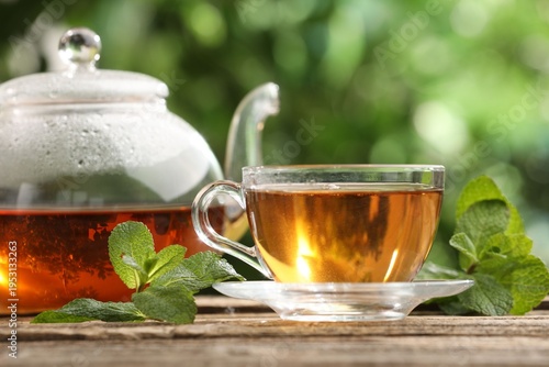 Fotografie Aromatic tea in glass cup, teapot and mint on wooden table outdoors, closeup