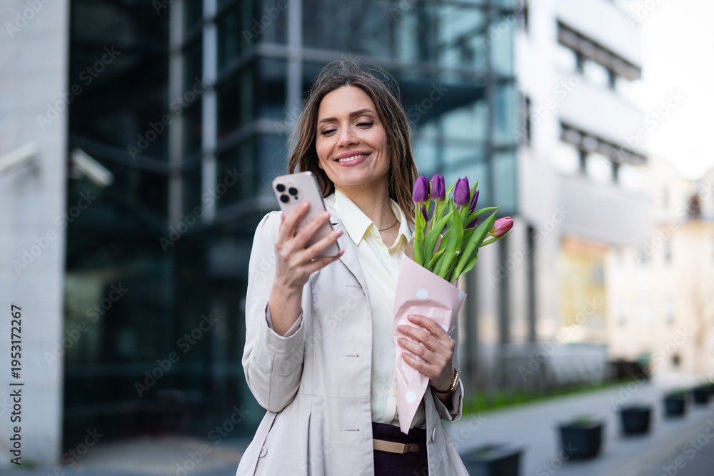 Fototapeta premium Smiling businesswoman holding a bouquet of tulips while looking at her smartphone outdoors