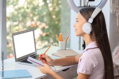 Teenage girl with headphones and laptop studying at table in bedroom