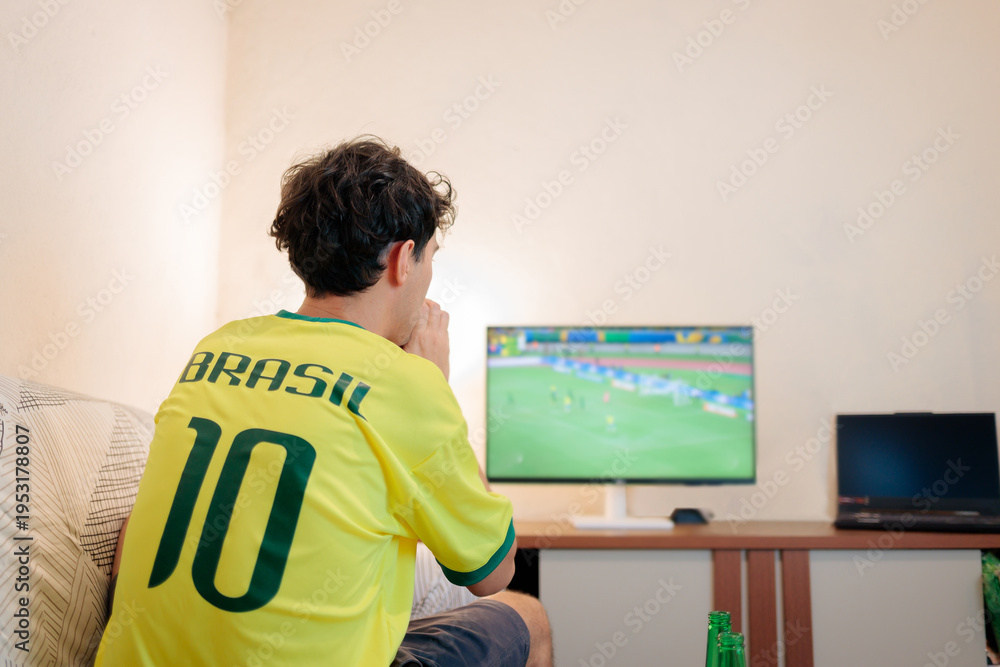 Fototapeta premium Torcedor brasileiro assistindo a uma partida internacional de futebol em casa, visto de costas com a camisa da seleção. Jovem focado na TV em momento decisivo, transmitindo tensão e expectativa.