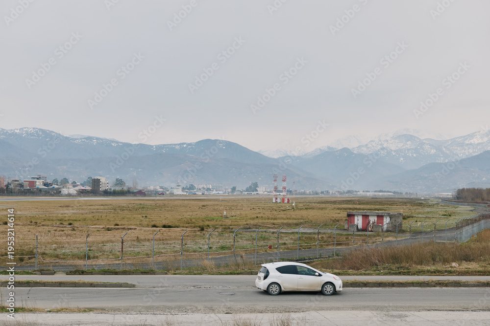 Fototapeta premium White compact car parked on roadside near field with distant mountains under cloudy sky. Concept of travel, transportation, countryside, and nature landscape in calm weather conditions.