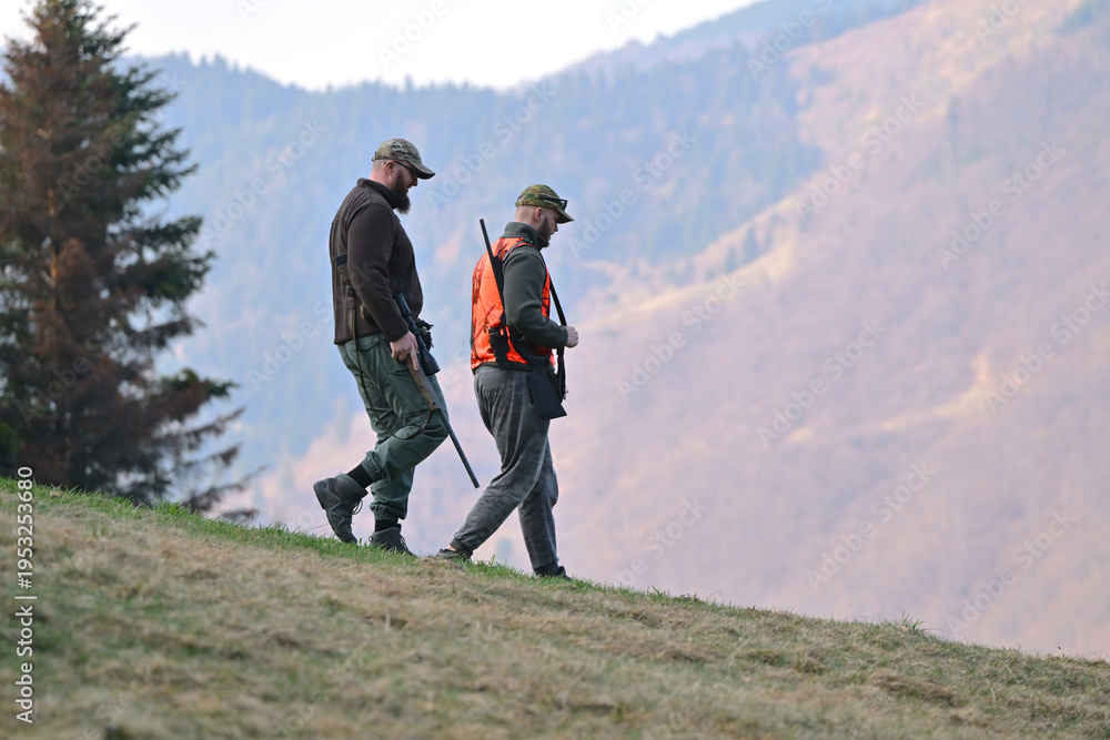 Obraz premium Two hunters walking down a mountain slope carrying rifles during autumn hunting trip