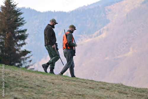 Two hunters walking down a mountain slope carrying rifles during autumn hunting trip